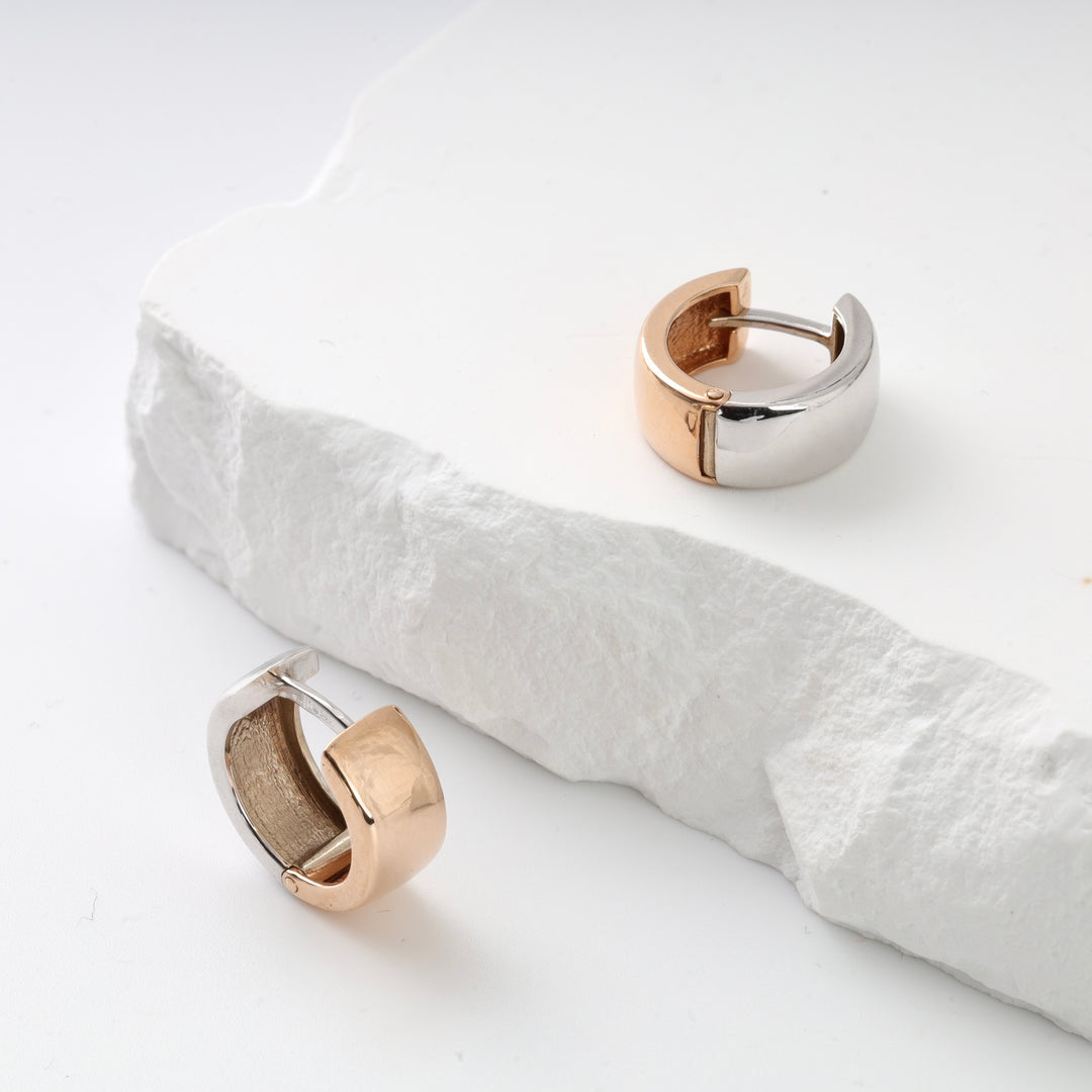 Pair of two-tone gold and silver chunky huggie hoop earrings displayed on a white stone surface.