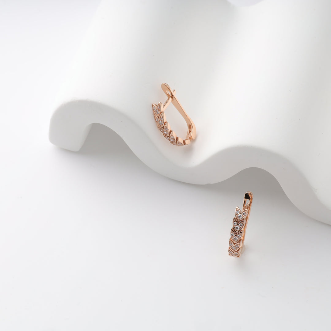 Rose gold huggie earrings with leaf motif and cubic zirconia on a white wavy surface.