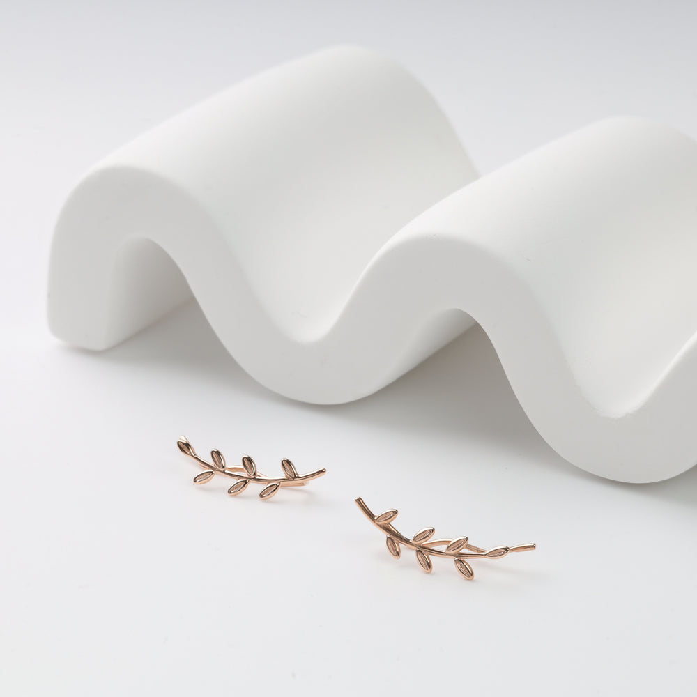 Rose gold leaf ear climber earrings, pair of metal vine studs displayed on a white wavy prop.