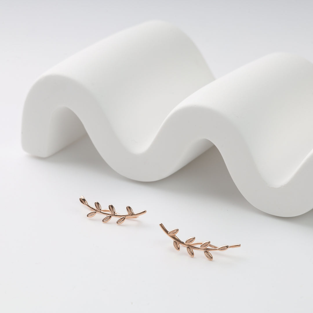Rose gold leaf ear climber earrings, pair of metal vine studs displayed on a white wavy prop.