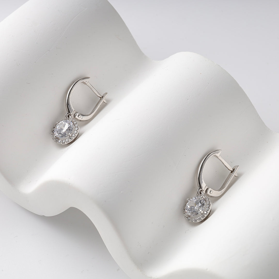 Pair of silver drop earrings with clear oval crystal center and small stones, displayed on a wavy white surface.