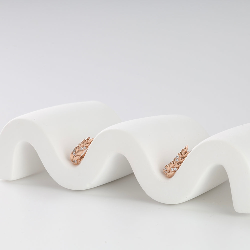 Pair of rose gold leaf motif hoop earrings with small clear stones displayed on a white wavy stand.