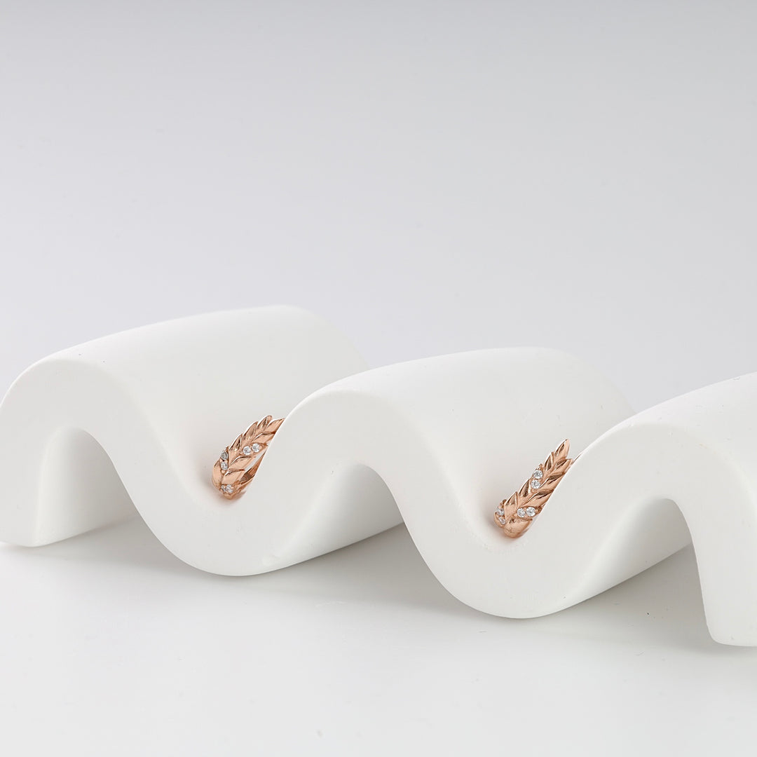 Pair of rose gold leaf motif hoop earrings with small clear stones displayed on a white wavy stand.