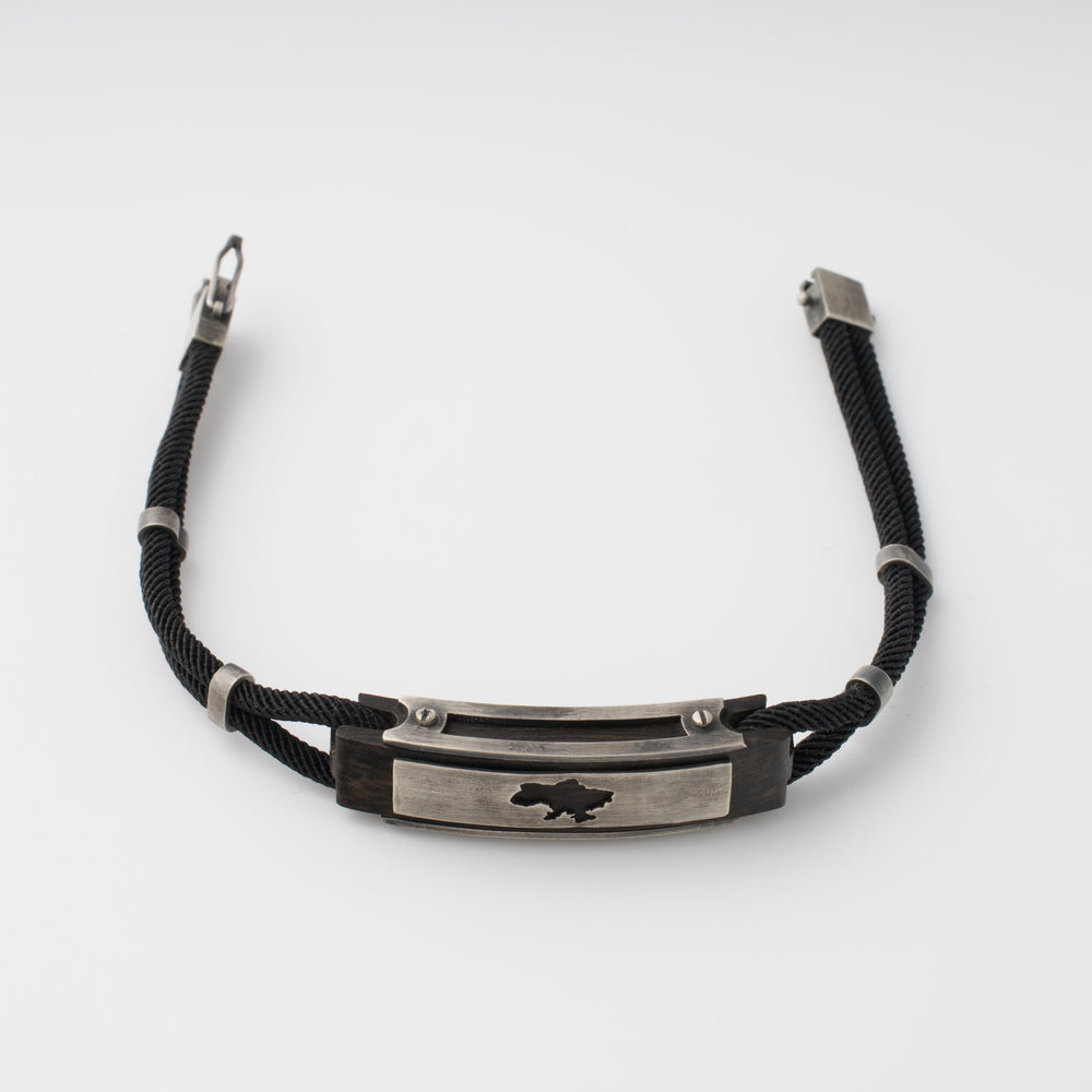 Black cord bracelet with a silver metal plate featuring a cutout map motif, displayed on a white background.