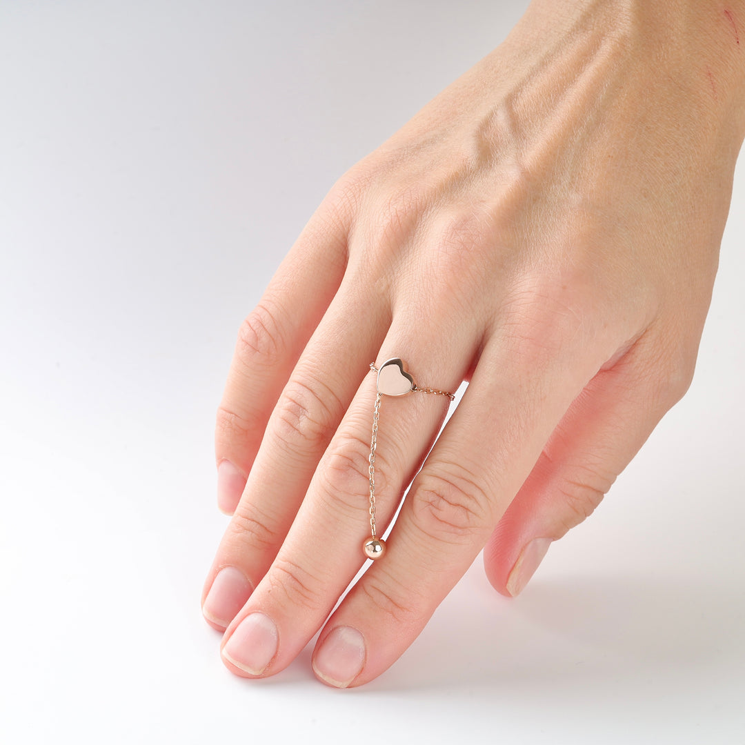Gold chain ring with a heart charm and dangling bead worn on a hand, photographed on a white background.