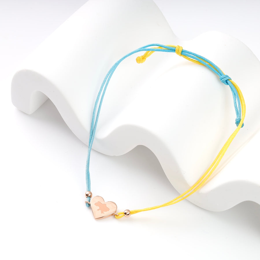 Blue and yellow string bracelet with rose gold heart charm displayed on wavy white surface.