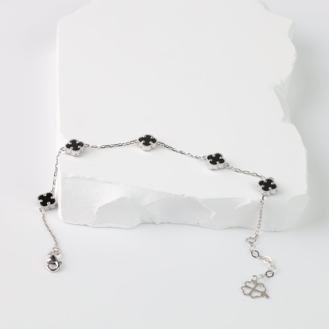 Silver chain bracelet with five black enamel clover charms displayed on a white stone background.