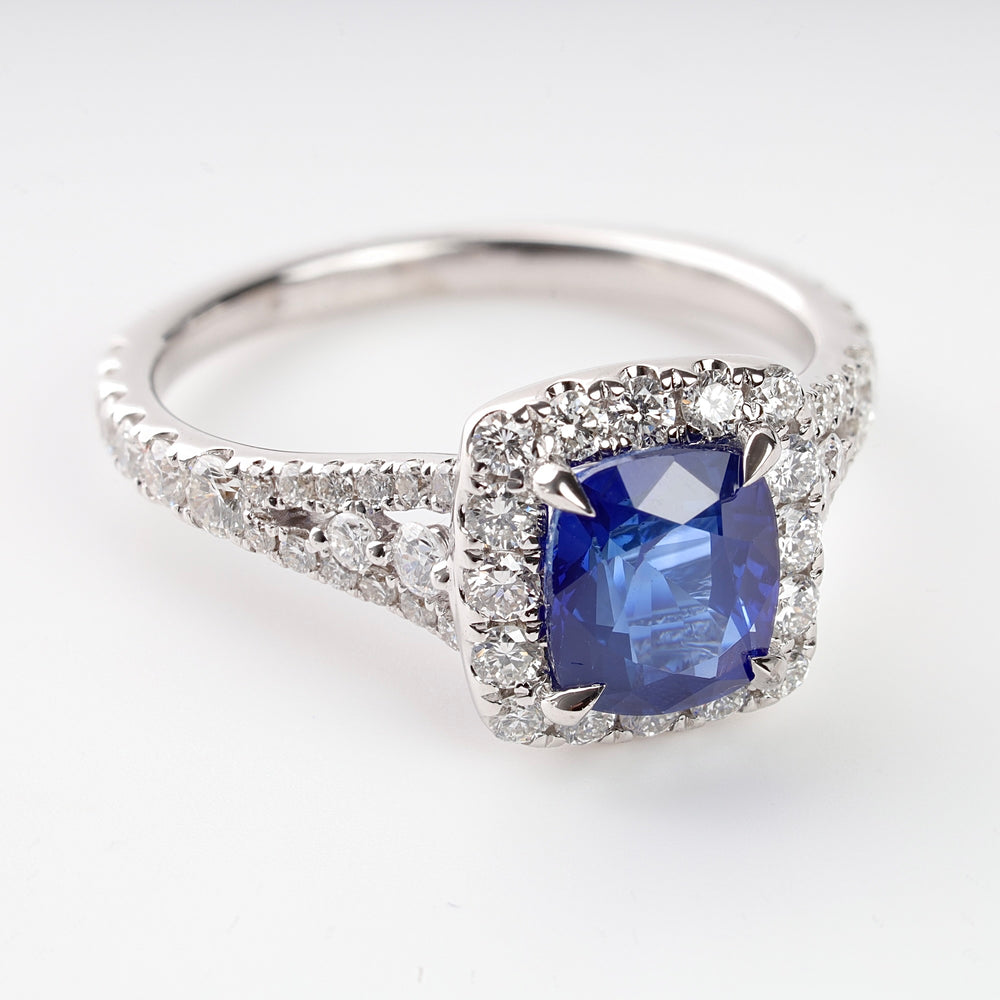 White gold ring with a cushion-cut blue sapphire center and halo of round white diamonds on a white background.