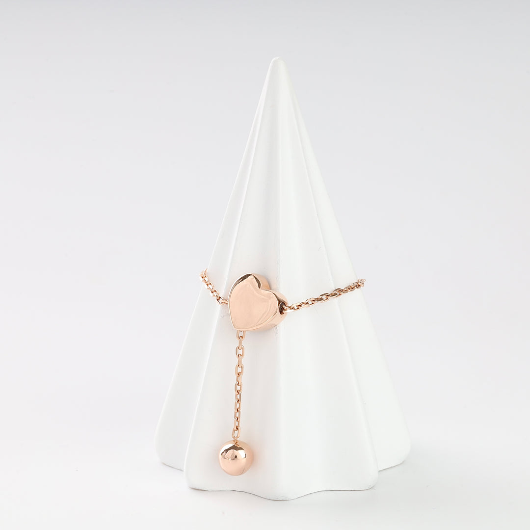 Rose gold chain ring with a heart charm and dangling ball, displayed on a white cone stand.