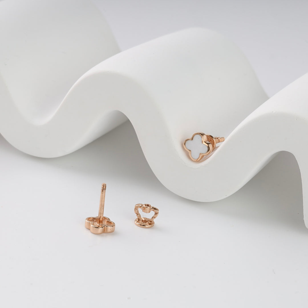 Pair of rose gold clover stud earrings with white enamel detail displayed on a white wavy stand.