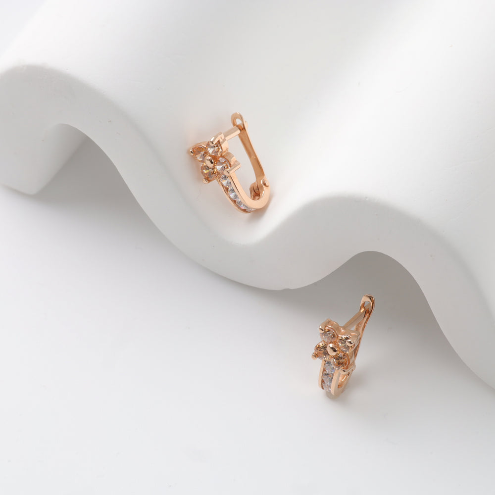 Rose gold huggie earrings with clear crystals and floral motif displayed on white curved surface.