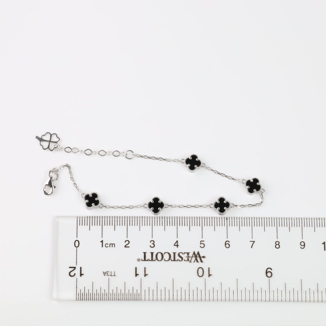 Silver chain bracelet with five black enamel clover charms, shown next to a clear plastic ruler on a white background.