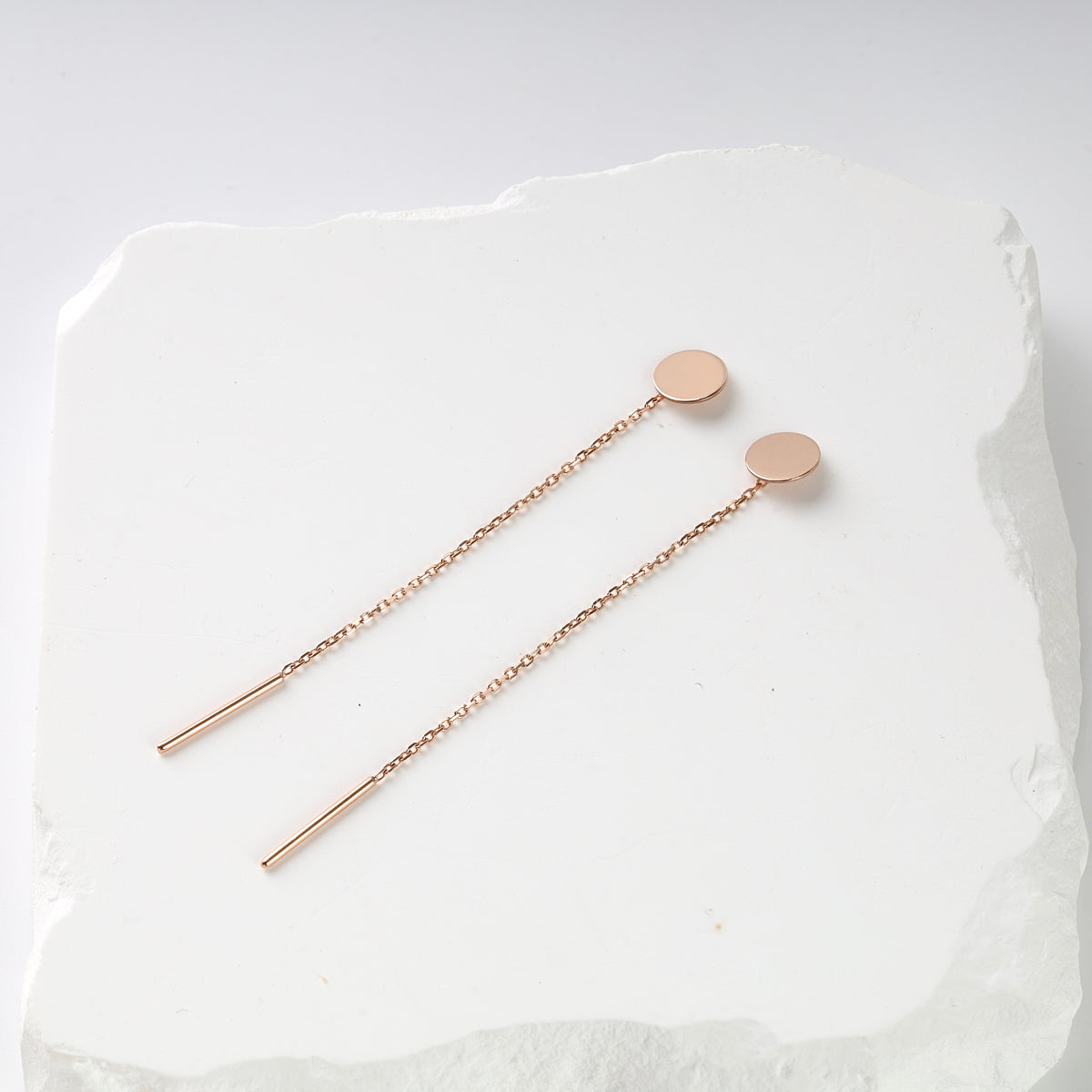 Pair of rose gold threader earrings with round disc tops and long chain drops displayed on a white textured surface.