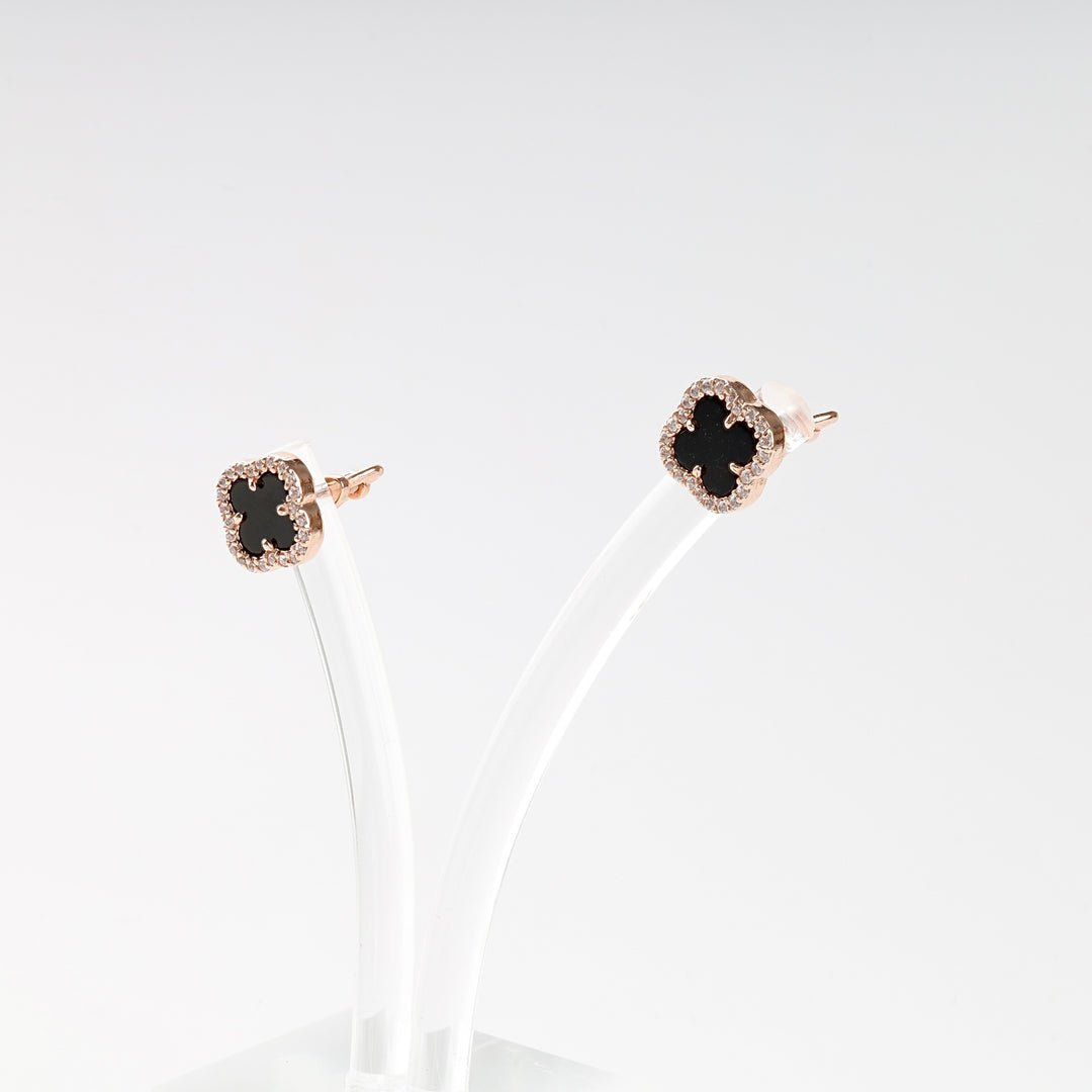 Pair of rose gold stud earrings with black clover-shaped stones and sparkling trim, displayed on a clear stand.