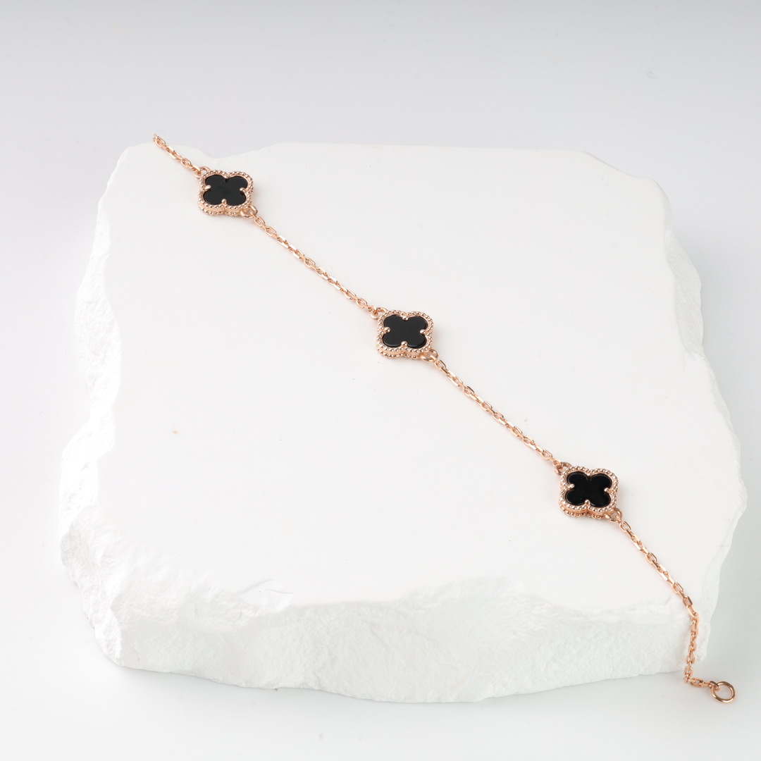 Rose gold chain bracelet with three black clover charms displayed on a white stone slab.