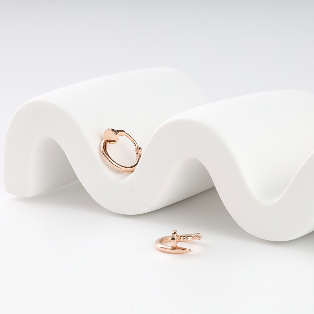 A pair of rose gold huggie earrings shaped like nails, displayed on a white wavy surface.