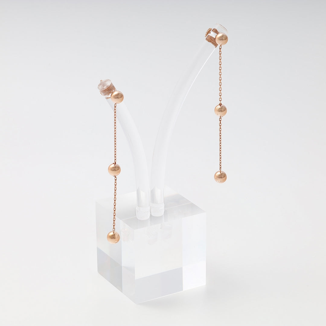 Pair of rose gold drop earrings with three polished ball beads on slender chains, displayed on a clear acrylic stand.