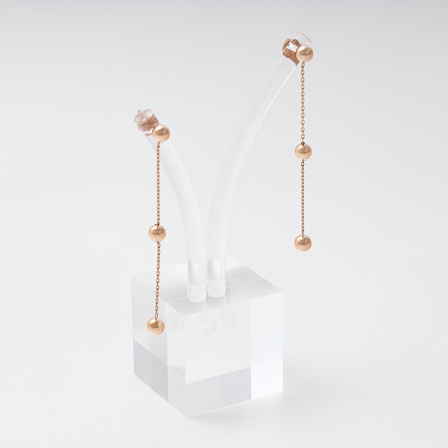 Pair of rose gold drop earrings with three polished ball beads on slender chains, displayed on a clear acrylic stand.