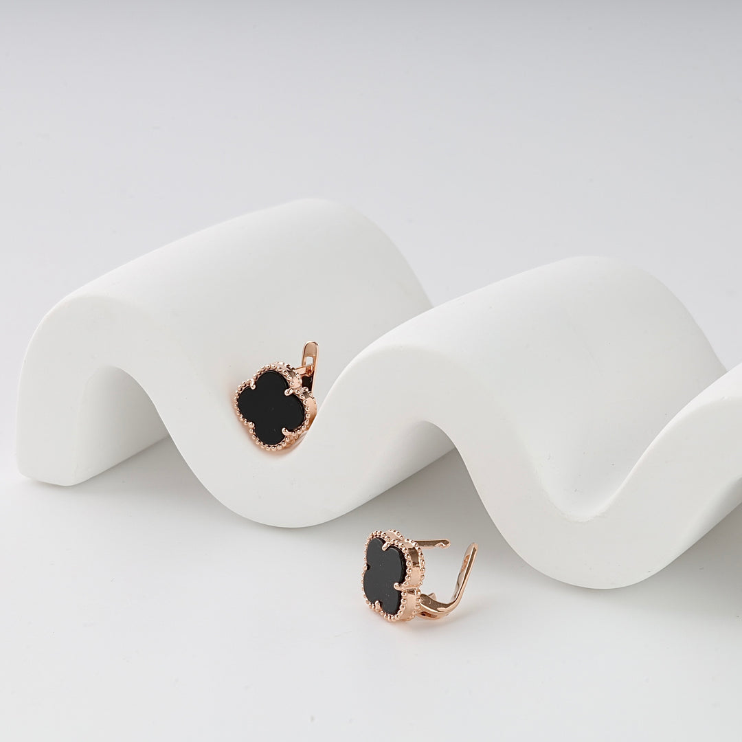 Pair of gold clover-shaped black enamel earrings displayed on a curved white stand.