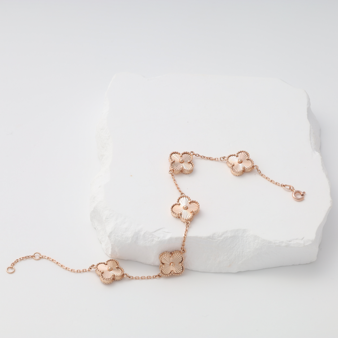 Rose gold chain bracelet with five clover flower charms displayed on a white stone surface.