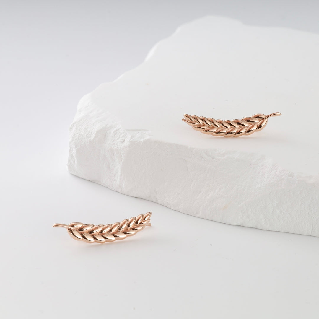 Two rose gold leaf-shaped earrings with braided detail displayed on a white textured stone surface.