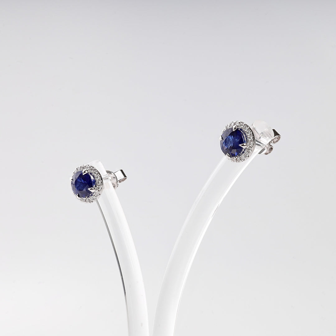 Pair of round silver stud earrings with blue gemstone centers and clear crystal halo, displayed on a white stand.