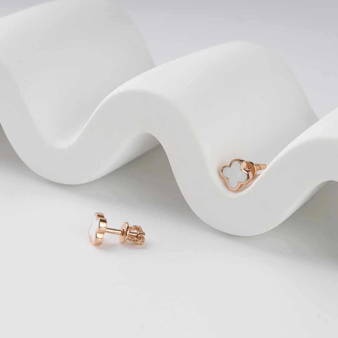 Pair of rose gold clover stud earrings with white enamel detail displayed on a white wavy ceramic surface.