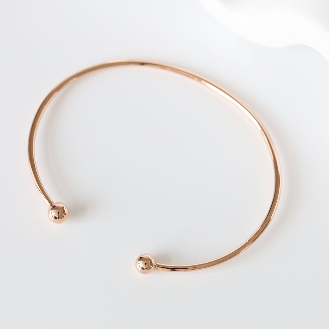 Rose gold open bangle bracelet with ball ends displayed on a white background, minimalist jewelry, cuff bracelet.
