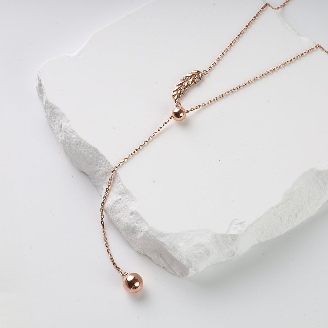 Rose gold lariat necklace with a leaf charm and two polished beads displayed on a white stone background.