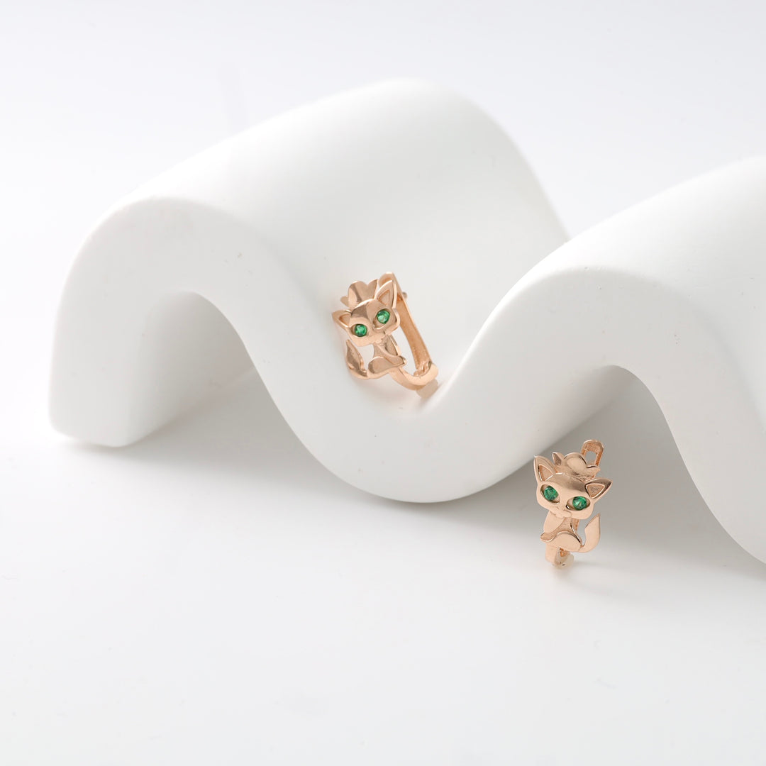 Rose gold cat earrings with green eyes on a white wavy display.