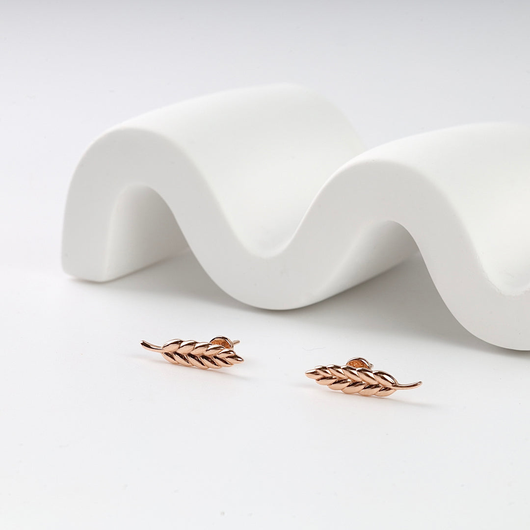 Pair of rose gold leaf stud earrings displayed on a white wavy background, botanical jewelry, minimalist earrings.