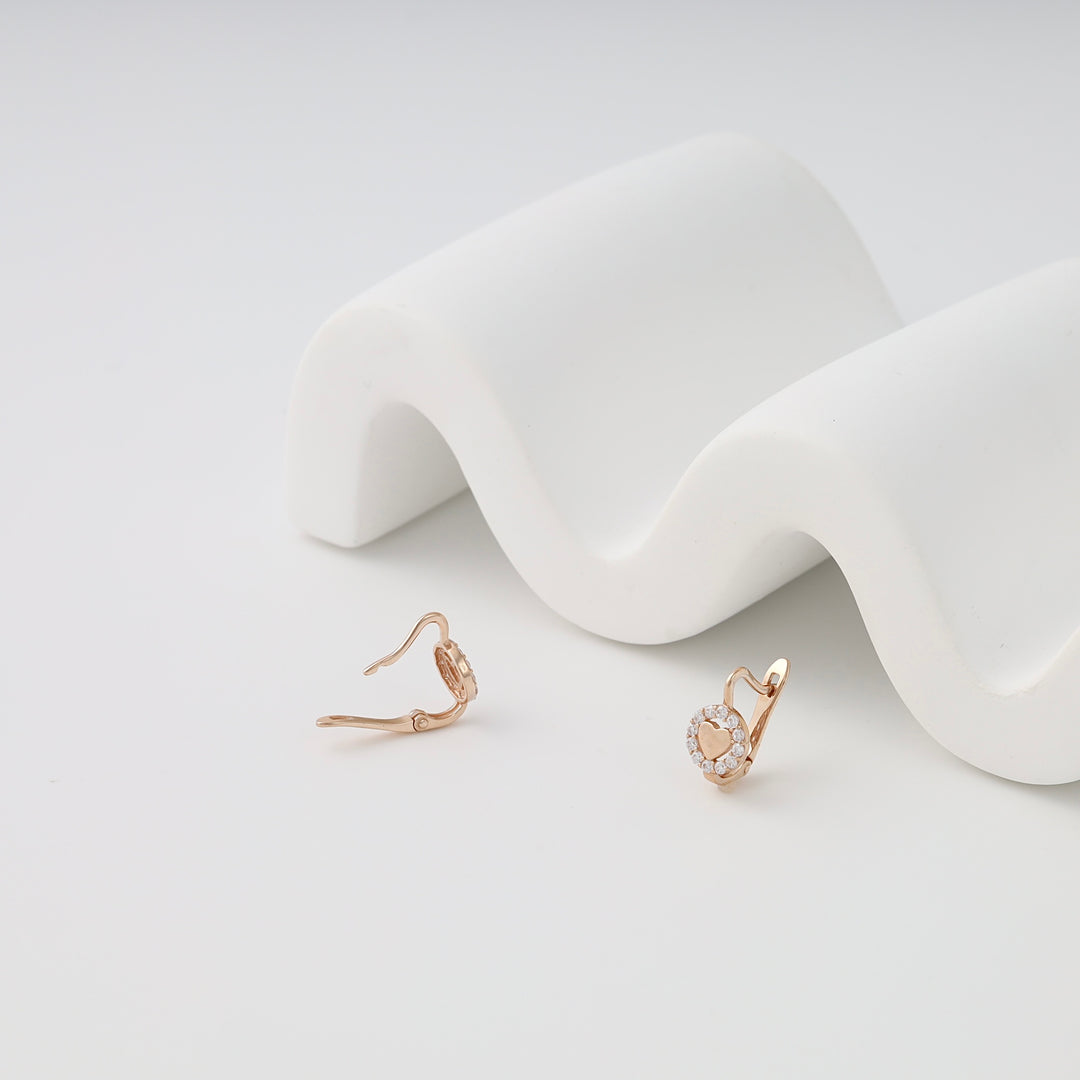 Pair of rose gold leverback earrings with a heart motif and clear crystals, shown on a white wavy background, dainty jewelry.