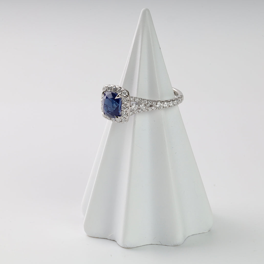 Silver ring with a square blue gemstone and clear stone accents displayed on a white cone stand. Sapphire engagement ring.