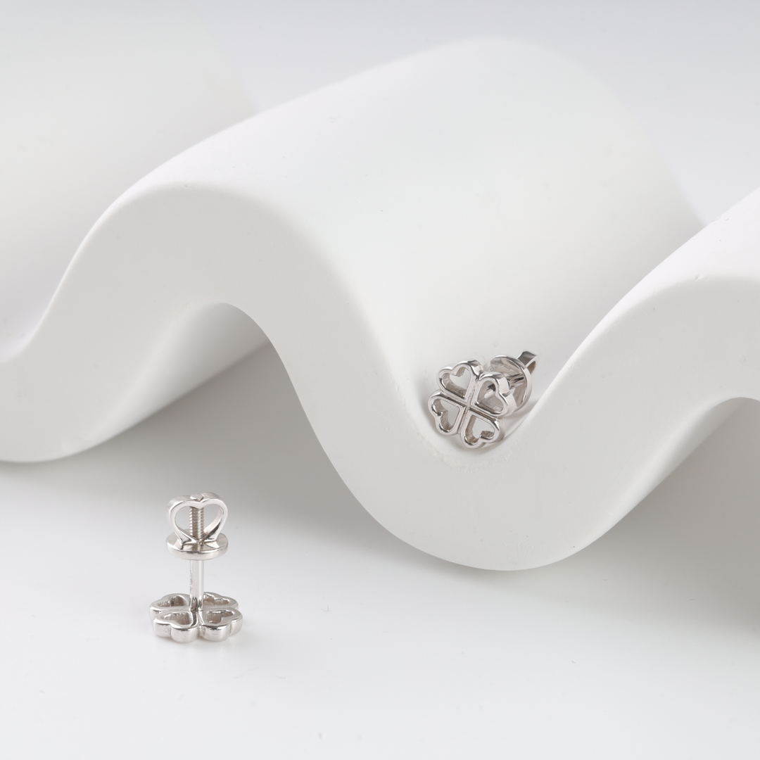 Pair of silver heart motif clover stud earrings displayed on a white wavy surface.