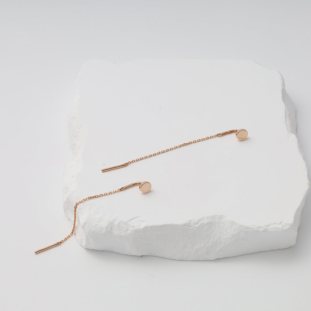 Pair of rose gold threader earrings with round discs and fine chains displayed on a white textured stone.