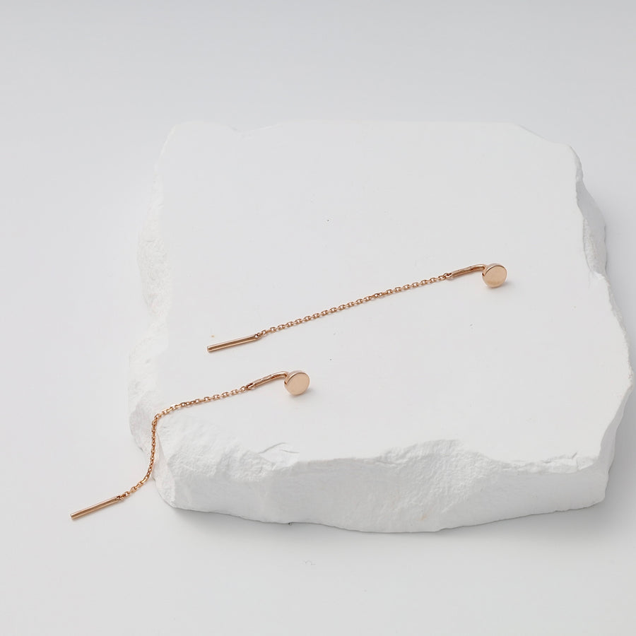 Pair of rose gold threader earrings with round discs and fine chains displayed on a white textured stone.