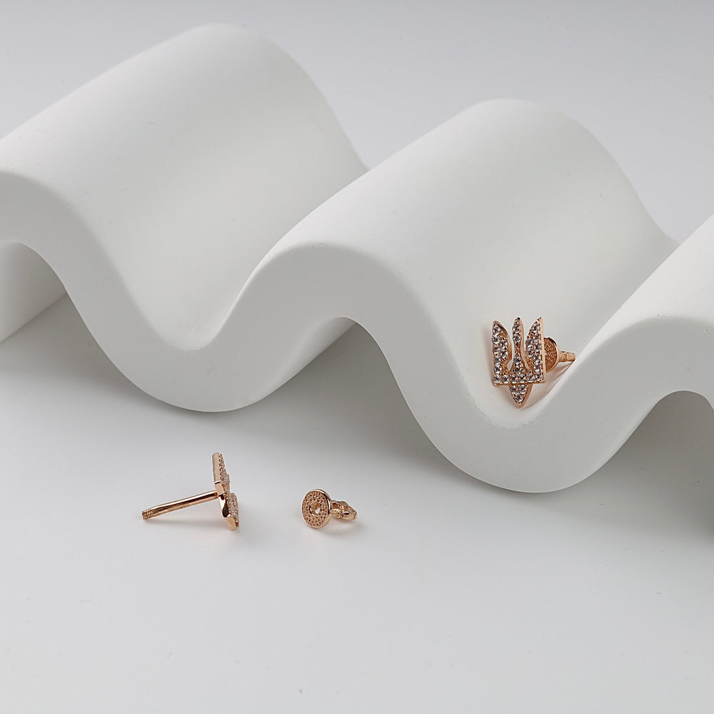 Rose gold stud earrings featuring pavé crystals and a trident motif, displayed on a white wavy surface.