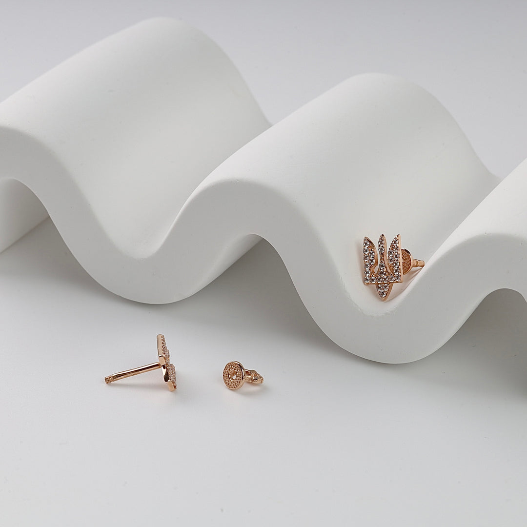 Rose gold stud earrings featuring pavé crystals and a trident motif, displayed on a white wavy surface.