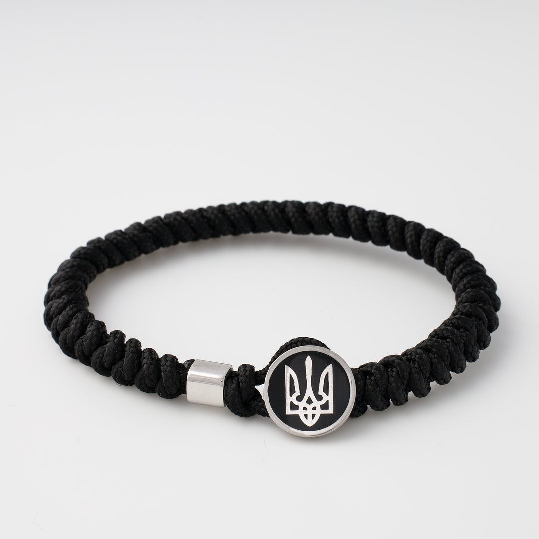 Black paracord bracelet with a round metal charm featuring the Ukrainian trident symbol, shown on a white background.