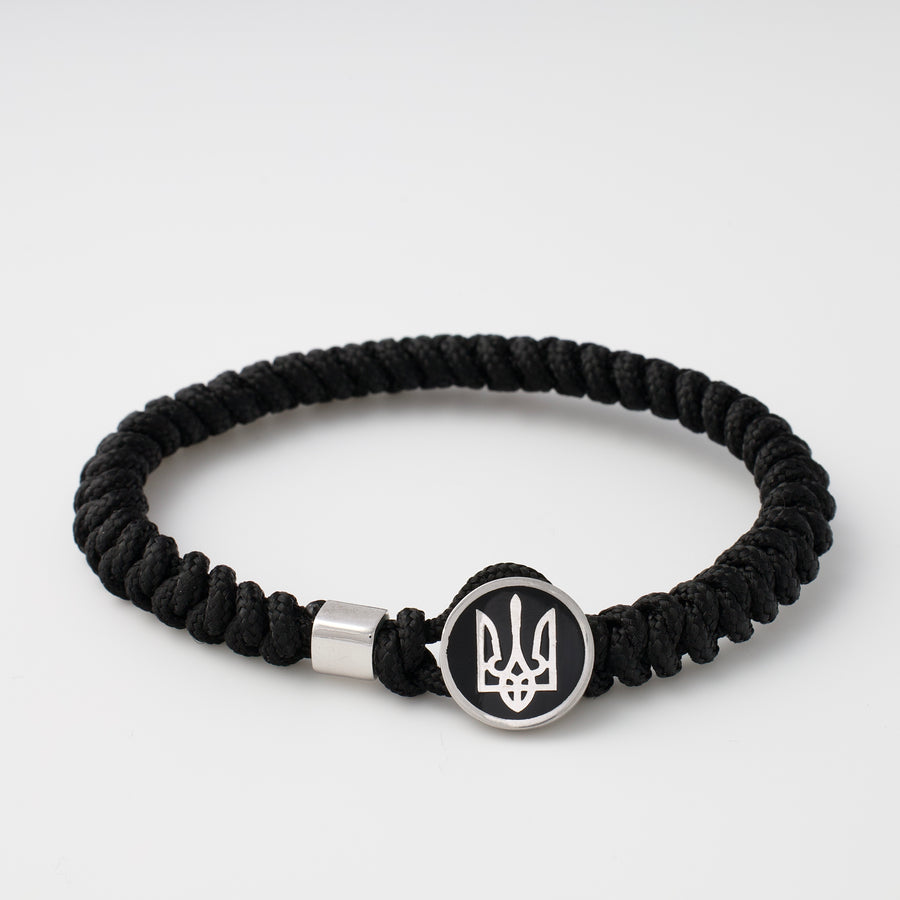 Black paracord bracelet with a round metal charm featuring the Ukrainian trident symbol, shown on a white background.