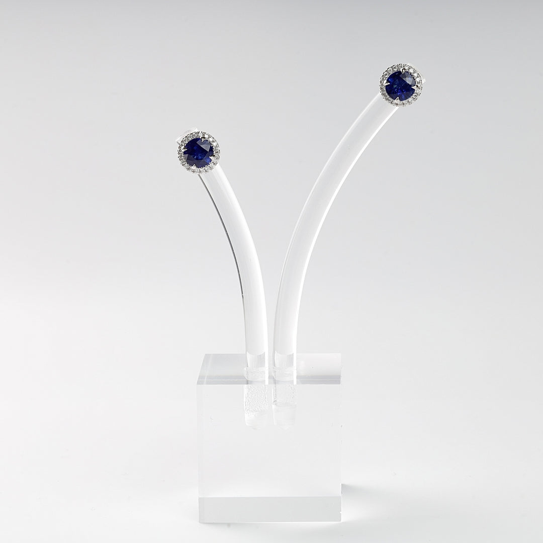 Pair of round blue sapphire stud earrings with a halo of clear stones, displayed on a white acrylic stand.