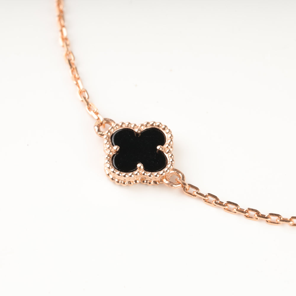 Rose gold chain bracelet with a black enamel quatrefoil charm set in a textured bezel, close up on white background.
