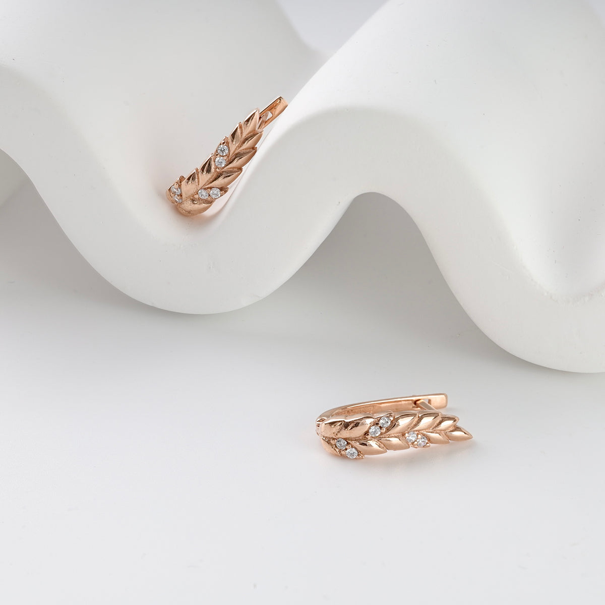 Rose gold earrings with leaf design and small clear crystals, displayed on a white curved background.