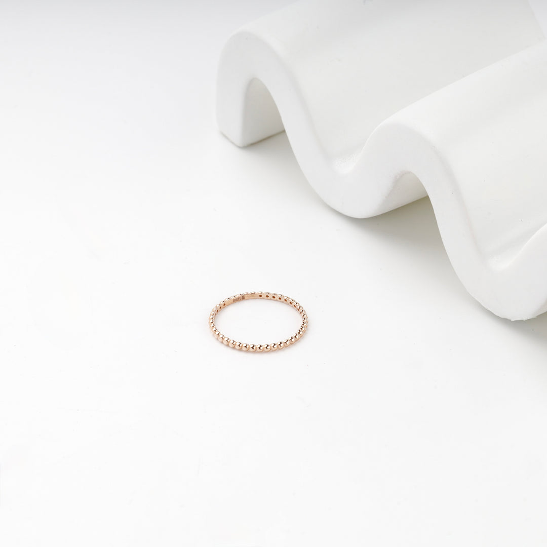 Rose gold beaded band ring on a white surface beside a wave-shaped stand