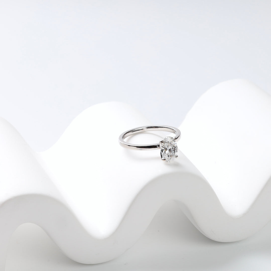 14K white gold oval lab-grown diamond solitaire ring resting on a white wave-shaped stand, side view.