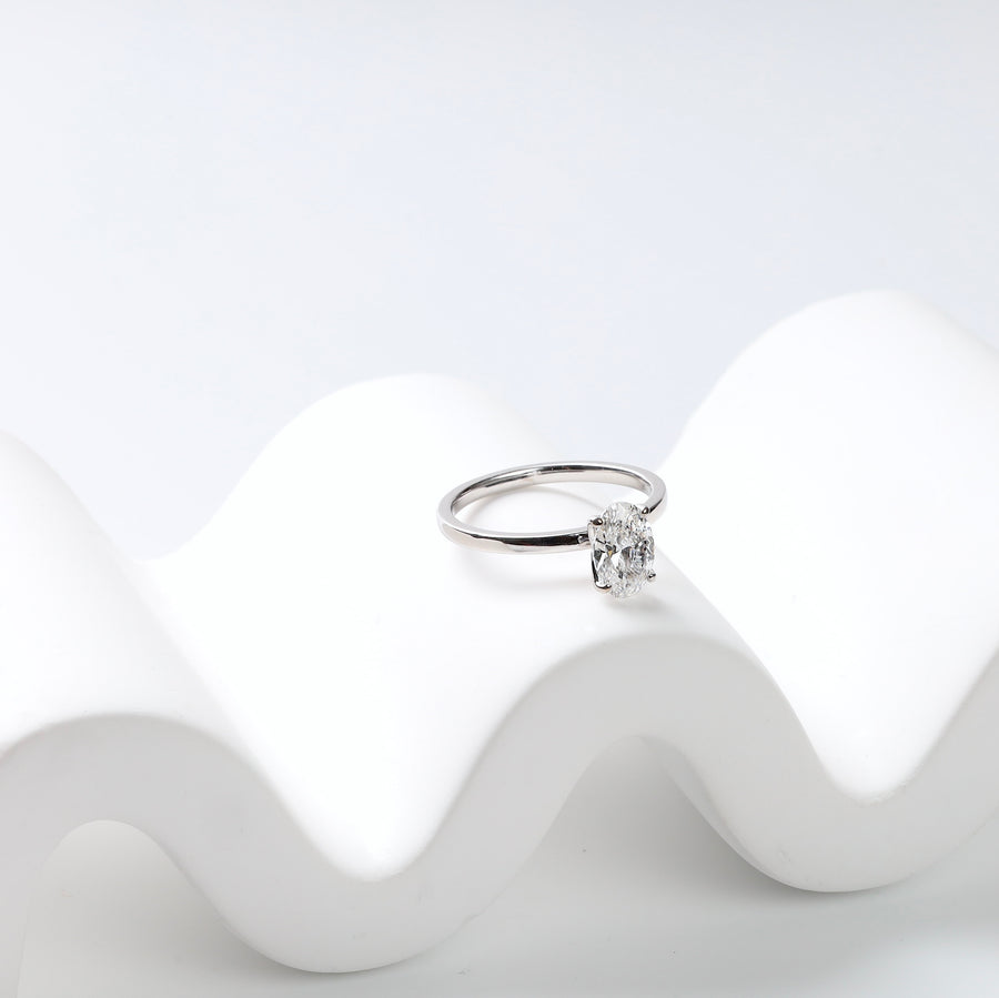 14K white gold oval lab-grown diamond solitaire ring resting on a white wave-shaped stand, side view.