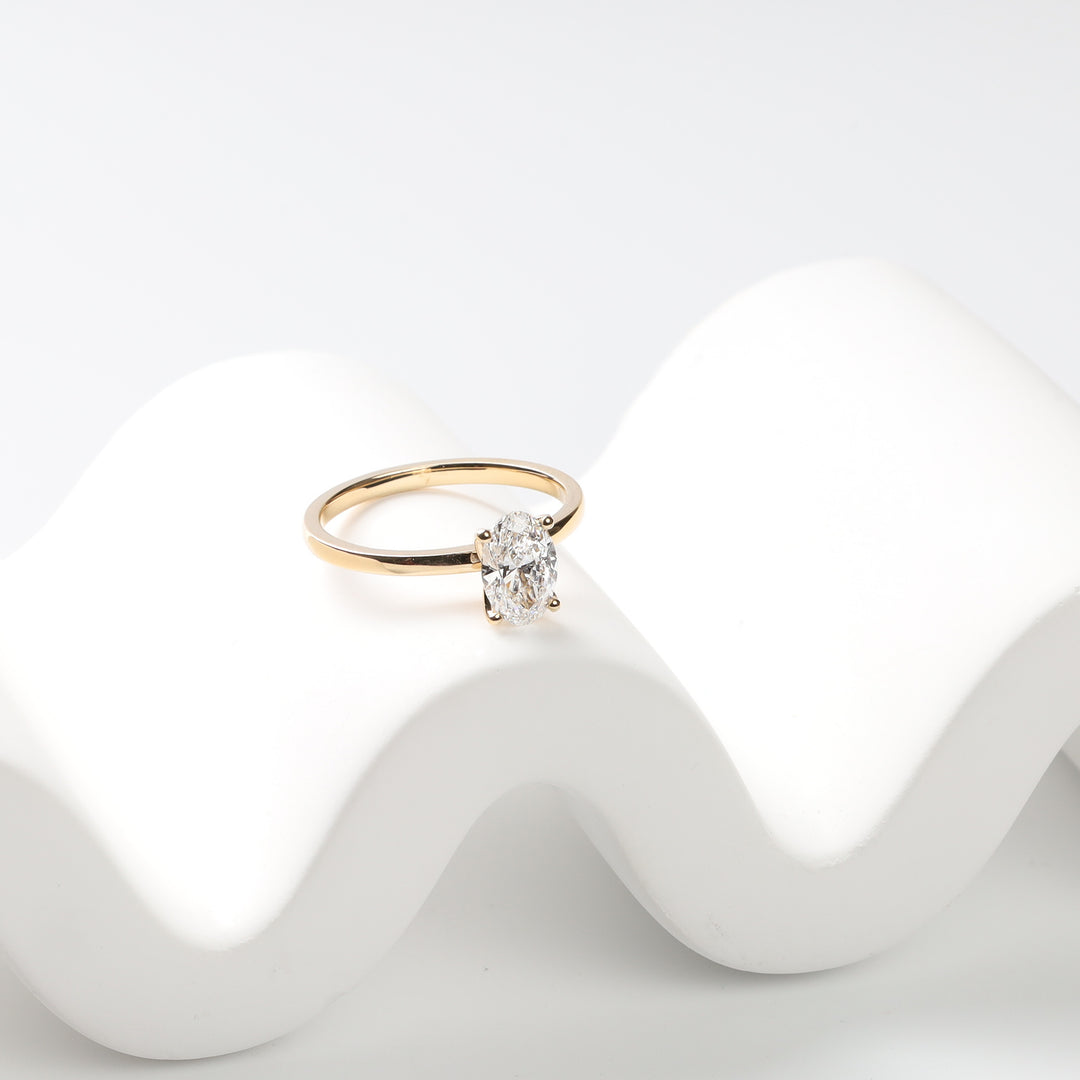 18K yellow gold oval lab-grown diamond solitaire ring on a white wave-shaped stand.