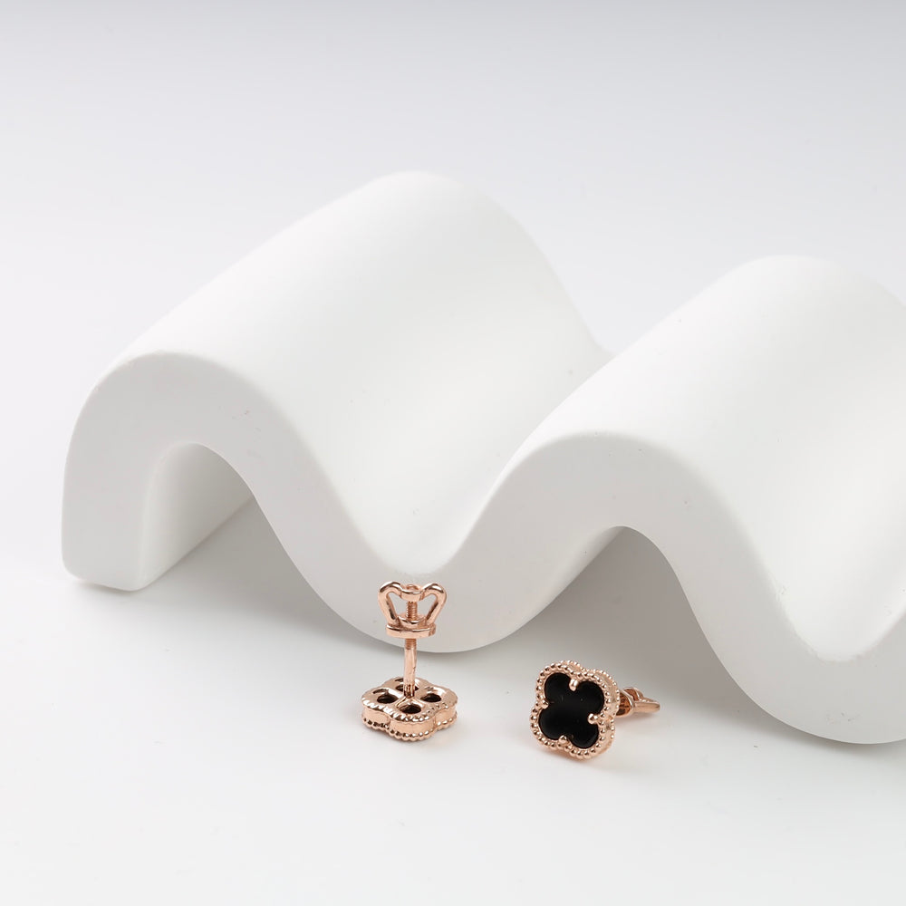 Rose gold and black enamel clover stud earrings on a wavy white surface.