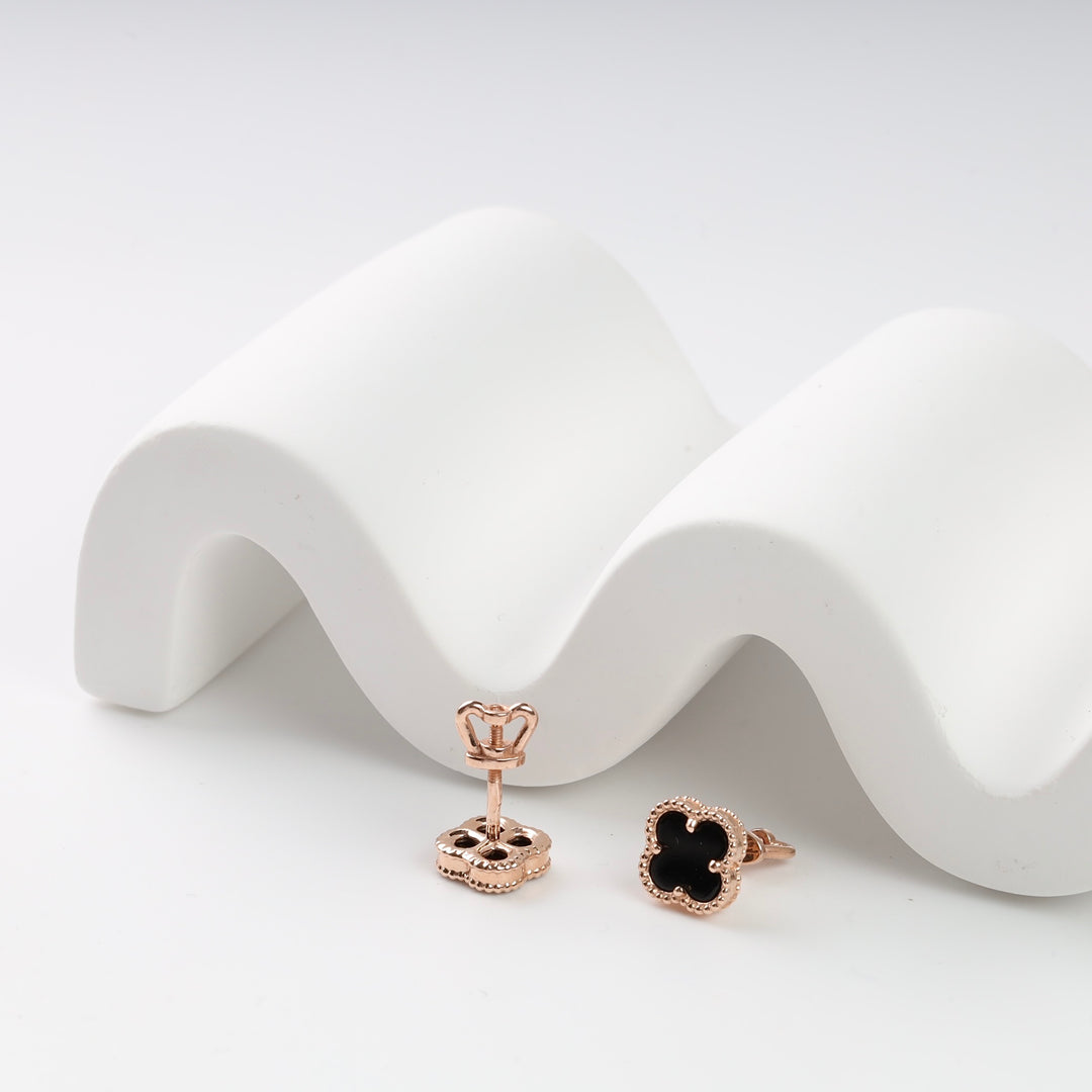 Rose gold and black enamel clover stud earrings on a wavy white surface.