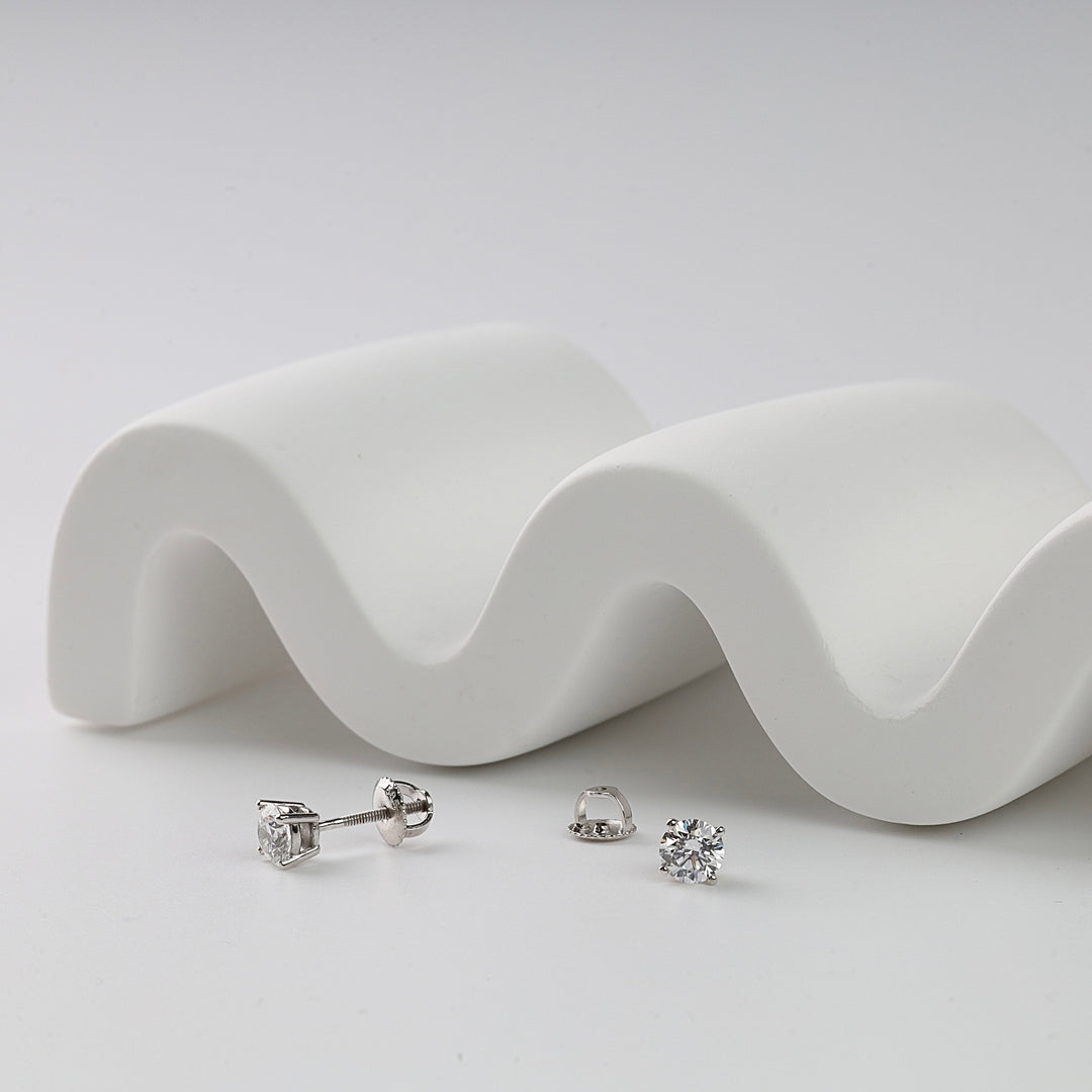 Pair of round diamond stud earrings with white gold settings displayed on a white surface with a wavy prop.